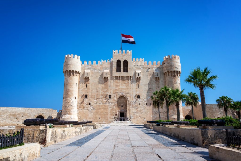 ALEXANDRIA Historic fortress with palm trees and a flag against a clear blue sky.