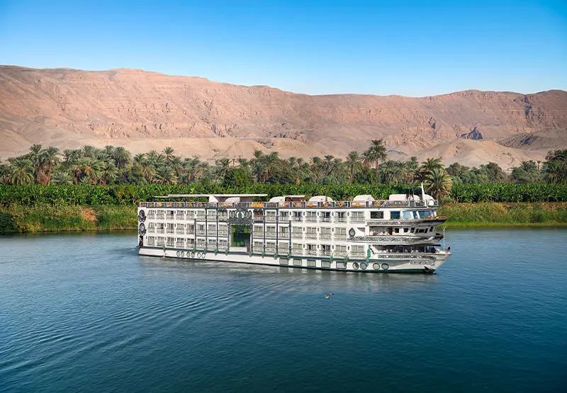 NILE CRUISES A modern cruise ship sailing on a river, surrounded by palm trees and mountains.